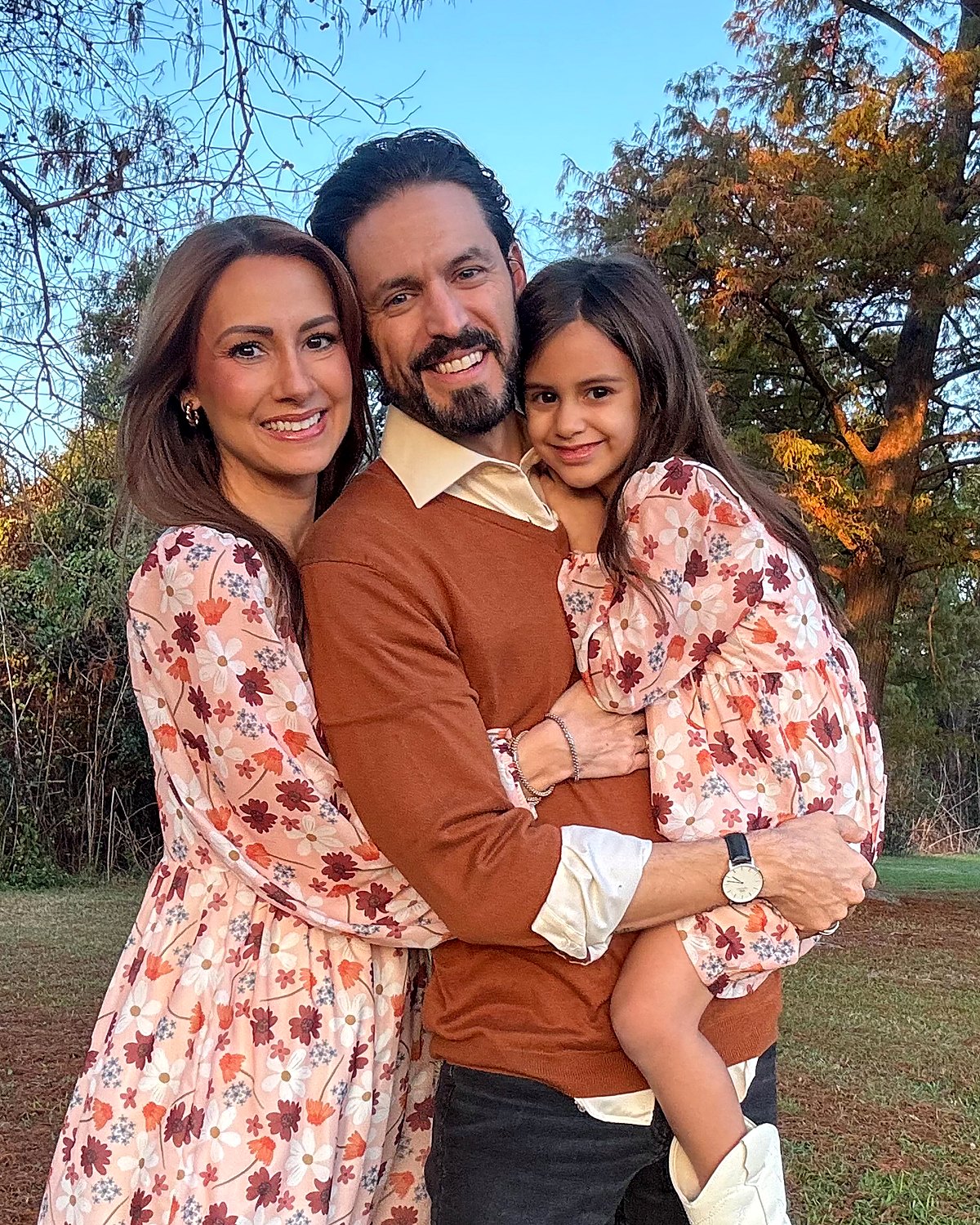 Dean and Michelle West outdoors in autumn, Dean holding their daughter between them, all three smiling at the camera.
